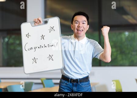 A young teenage male student is holding a sign saying congratulations on a whiteboard, with his arm making a fist pump gesture. He is in a classroom. Stock Photo