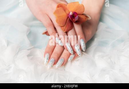 A close-up capture reveals a persons hand with intricate floral nail art delicately cradling an orange blossom. The surrounding soft, billowy texture Stock Photo