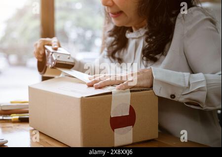 A cropped image of a mature Asian female clothes online seller packing a shipping cardboard box to ship to her customers, using a packing tape dispens Stock Photo