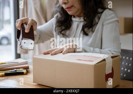 A cropped image of a mature Asian female clothes online seller packing a shipping cardboard box to ship to her customers, using a packing tape dispens Stock Photo