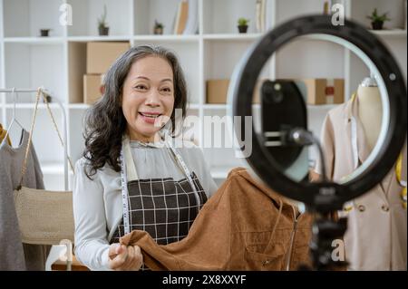 An attractive mature Asian female online seller is showing her goods live on her social media, talking with her customers on streaming, and selling cl Stock Photo