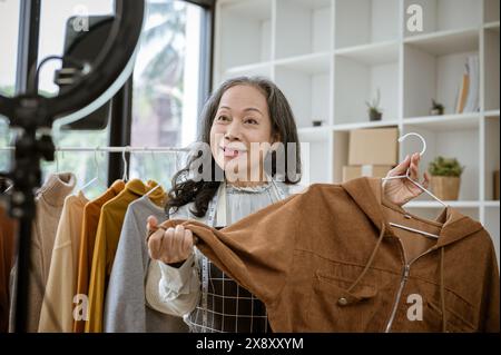 An attractive mature Asian female online seller is showing her goods live on her social media, talking with her customers on streaming, and selling cl Stock Photo