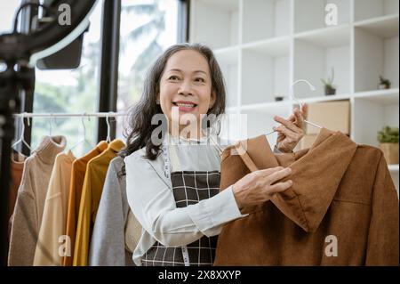 An attractive mature Asian female online seller is showing her goods live on her social media, talking with her customers on streaming, and selling cl Stock Photo