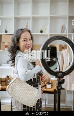 An attractive mature Asian female online seller is live streaming from her home office, selling her products online via social media, or recording a p Stock Photo