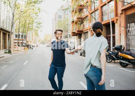 Couple walks hand in hand down a city street, smiling and looking at each other, enjoying a sunny day. Stock Photo