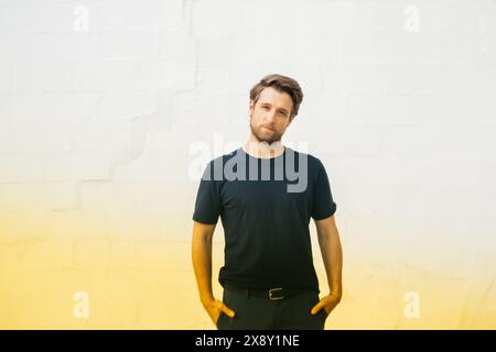Portrait of a man standing casually with his hands in his pockets, wearing a dark shirt and jeans, against a white brick wall background. Stock Photo