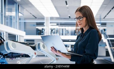 Beautiful Biotechnology Engineer Using Laptop Computer, Analyzing Robotic Machine Concept in a High Tech Factory. Female Scientist Manipulate and Program the Robot for Work. Stock Photo