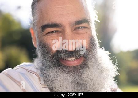 Caucasian senior man with grey beard, wearing light striped shirt, smiling outdoors Stock Photo