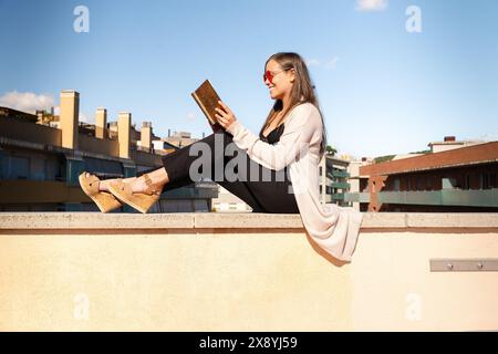Happy young woman sitting on the roof reading a book outdoors Stock Photo
