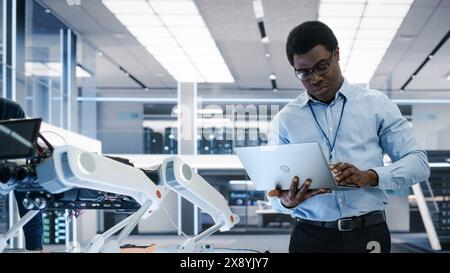 Smart Electronics Engineer Using Laptop Computer, Analyzing Robotic Machine Concept in a High Tech Factory. African Scientist Manipulate and Program the Robot for Work. Stock Photo