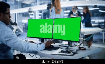 Software Developer Working on Computer with Green Screen Mock Up Display. Scientific Lab, Engineering Research Center with Specialists Working on Mobile Robot Development in the Background. Stock Photo