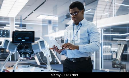 Handsome Biotechnology Engineer Using Laptop Computer, Analyzing Robotic Machine Concept in a High Tech Factory. African Scientist Manipulate and Program the Robot for Work. Stock Photo
