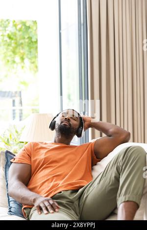 At home, African American man relaxing on couch, enjoying music, copy space. Wearing headphones, an orange shirt, and green pants, he has short black Stock Photo