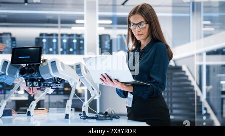Portrait of a Young Robotics Engineer Using Laptop Computer, Analyzing Robotic Machine Concept in a High Tech Factory. Female Scientist Manipulate and Program the Robot for Work. Stock Photo