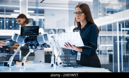 Portrait of a Young Robotics Engineer Using Laptop Computer, Analyzing Robotic Machine Concept in a High Tech Factory. Female Scientist Manipulate and Program the Robot for Work. Stock Photo