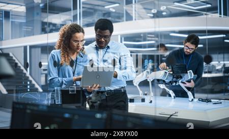 Portrait of Two Multiethnic Female and Male Engineers Use Laptop Computer to Connect, Analyze and Discuss How to Program a Robot Dog. Standing in High Tech Research Laboratory with Modern Equipment. Stock Photo