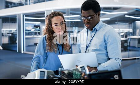 Two Young Multiethnic Engineers Use Laptop Computer to Analyze and Discuss How a Futuristic Robot Dog Works. High Tech Research Laboratory with Modern Equipment. Stock Photo