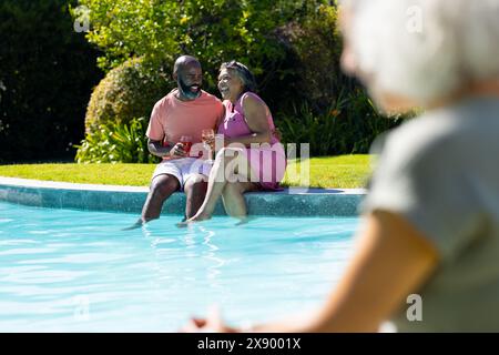 Outdoors, diverse senior friends, laughing by pool, holding drinks Stock Photo