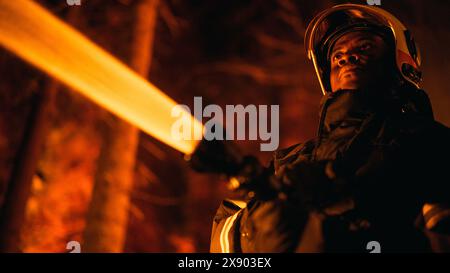 Close Up Low Angle Creative Portrait of a Brave African American Firefighter Using a Firehose to Fight a Forest Fire. Black Fireman Holds High-Pressure Water Hose in Hands And Saving Environment. Stock Photo