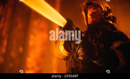 Close Up Low Angle Portrait of a Brave Professional Firefighter Using a Firehose to Fight a Raging Dangerous Forest Fire. Fireman Holds High-Pressure Water Hose in Hands and Battling Bushfire. Stock Photo