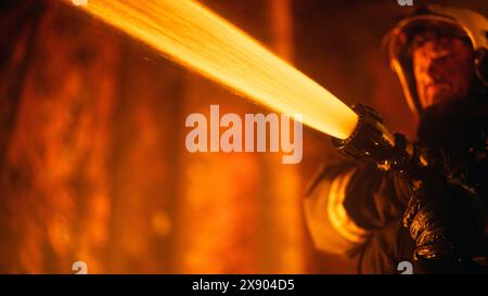 Close Up Low Angle Portrait of a Brave Professional Firefighter Using a Firehose to Fight a Raging Dangerous Forest Fire. Fireman Using High-Pressure Water Hose With Hands and Battling Bushfire. Stock Photo