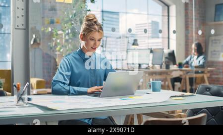 Portrait of Beautiful Caucasian Woman Working on Laptop Computer at a Bright Creative Office. Young Female Manager Updating Employees' Schedules and Answering Colleagues' E.mails Stock Photo