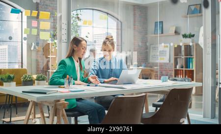 Two Colleagues Working On Laptop in Office. Female Help Desk Coordinator Collaborates with Caucasian Customer Service Agent, They Discuss a Project, Chat, Smile. Teamwork Collaboration Stock Photo
