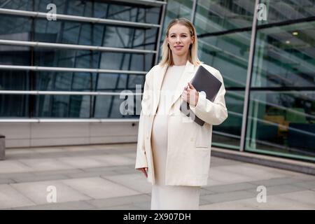 Beautiful pregnant blonde woman in a white business suit with a blazer stands outdoors with a laptop near a corporate building, showcasing professiona Stock Photo