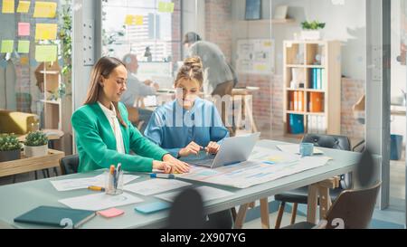 Portrait of Two Caucasian Smiling Women Collaborating and Working on a Laptop Computer in a Bright Office. Two Female Data Analysts Developing a Project and Preparing a Presentation. Teamwork Concept Stock Photo
