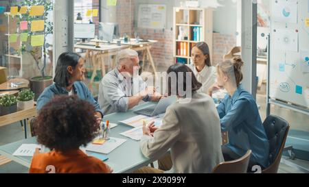In the Meeting Room Team of Entrepreneurs and Specialists sitting at the Conference Table Have Discussions about Statistics and Charts, Solve Problems, Led by Senior Lead. They are Using Laptop Stock Photo