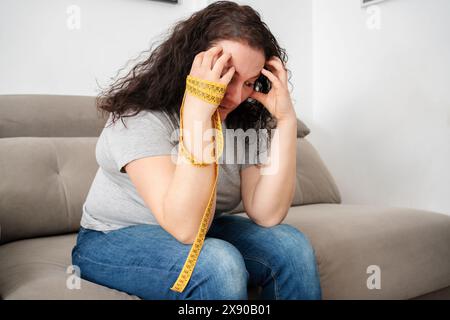 Sad woman worried about appearance holding a measure tape sitting on a couch in the living room at home Stock Photo