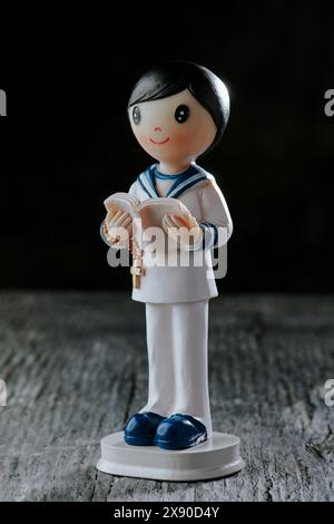 a figurine of a first communion boy, in a white sailor suit, wearing a rosary and a bible in his hands, standing on a rustic gray wooden surface Stock Photo