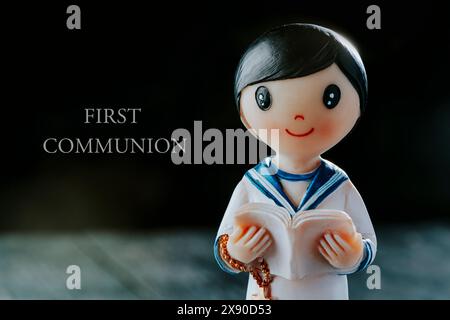 the text first communion and a figurine of a communicant boy, wearing a white sailor suit, a rosary and a bible, against a black background Stock Photo