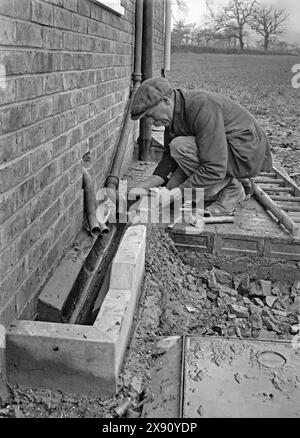 A building worker constructing an outside drainage gulley or channel. The work took place on a house in Kennel Lane, Fetcham, Leatherhead, Surrey, England, UK c. 1930. The pipework at this time included lead (the two pipes coming out through the wall) and cast iron (the larger downpipe from the roof guttering). The gulley itself is part concrete, with the worker finishing off the rear section in brick – a vintage 1920s/30s photograph. Stock Photo