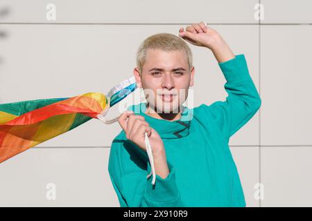 Portrait of a Young Latin Gay Man outdoors with a rainbow flag in his hand. He is smiling and looking at the camera. The flag is waving in the air Stock Photo