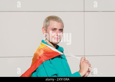 A Young Latin Man with short hair is holding a Pride Flag on his shoulders. He is is standing at dusk on the street, smiling looking at camera and he Stock Photo