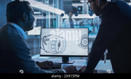 Two Turbine Engine Developers Working on Programming a Prototype Jet Engine on Desktop Computer. Young Team of International Industrial Science Specialists Developing a Turbine Motor in the Background Stock Photo