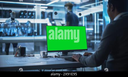 Software Developer Working on Computer with Green Screen Mock Up Display. Scientific Lab, Engineering Research Center with Specialists Working on Turbine Engine Development in the Background. Stock Photo