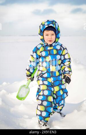A child dressed in a colorful snowsuit playing with a shovel in the snow on a cold winter day, Belarus, Minsk Stock Photo