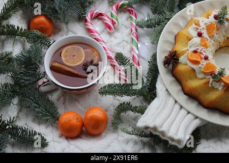 Cozy Christmas setting with a cup of spiced tea, candy canes, tangerines, and a cake with frosting on a white knitted background Stock Photo