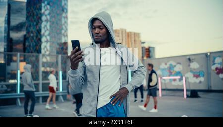 Handsome Black Man Holding a Smartphone. Young Stylish African Man Standing in a City Environment, Diverse People Playing Street Soccer in the Background Stock Photo