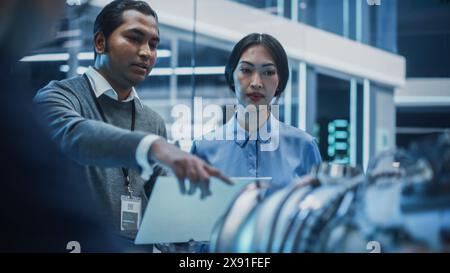 Two Multiethnic Employees Work in Modern Innovative Research Facility with a Futuristic Jet Engine. Engineer with a Laptop Explaining How the Prototype Motor Works to an Industrial Specialist. Stock Photo
