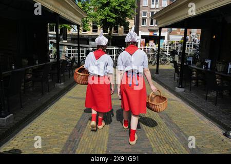 Two Dutch cheese girls in traditional tracht and carrying baskets of cheese, wander through the town of Alkmaar on market day., Alkmaar, The Stock Photo