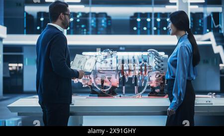Two Multiethnic Employees Standing in a Modern High Tech Research Facility with Futuristic Turbine Motor. Engineer Explaining How the Prototype Engine Works to an Industrial Specialist with a Laptop. Stock Photo