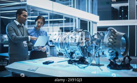 Two Multiethnic Employees Work in Modern Innovative Research Facility with a Futuristic Jet Engine. Engineer with a Laptop Explaining How the Prototype Motor Works to an Industrial Specialist. Stock Photo