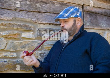 Close up of a man wearing a blue plaid cap smoking a red pipe while leaning against a rock wall. Stock Photo