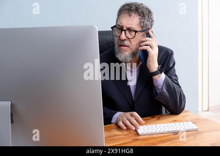 Professional businessman talking on the phone while working at a computer in his office - Corporate communication, professionalism, and efficient work Stock Photo