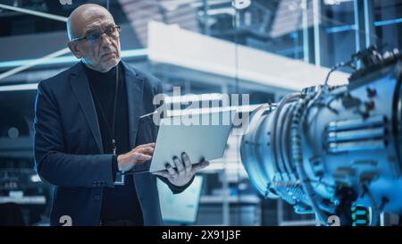 Middle Aged Industrial Engineer Working on a Futuristic Jet Engine, Standing with Laptop Computer in Scientific Technology Lab. Scientist Developing a New Electric Motor in a Research Facility. Stock Photo
