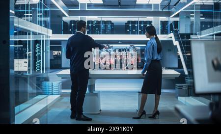 Two Diverse Engineers Working in High Tech Research Facility with a Futuristic Turbine Motor. Project Leader Explaining How the Prototype Engine Works to an Industrial Specialist with a Laptop. Stock Photo