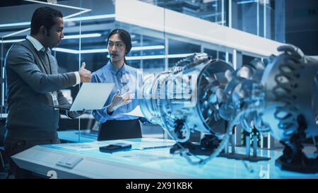 Two Multiethnic Employees Work in Modern Innovative Research Facility with a Futuristic Jet Engine. Engineer with a Laptop Explaining How the Prototype Motor Works to an Industrial Specialist. Stock Photo